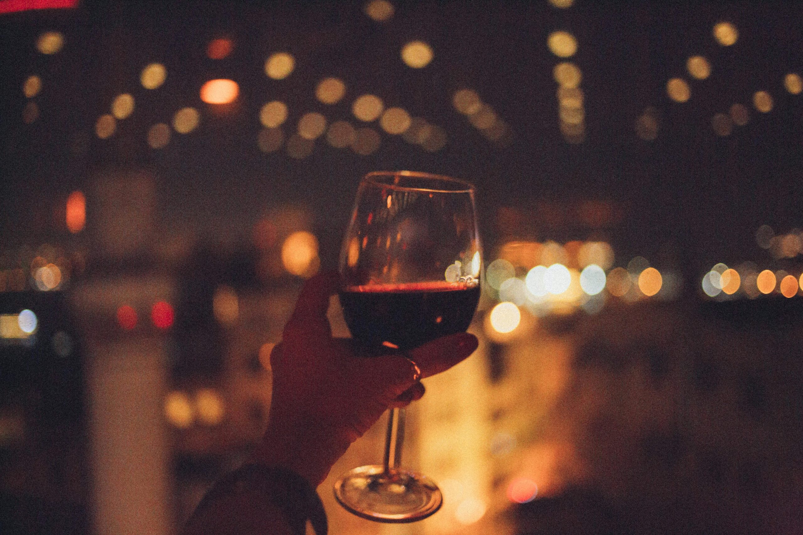 Accueil A hand holds a red wine glass with bokeh city lights in a nighttime setting.